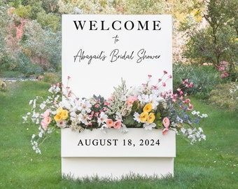 May include: White sign with the text "WELCOME to Abagail's Bridal Shower AUGUST 18, 2024" in black lettering. The sign is decorated with a floral arrangement of white, pink, and yellow flowers.