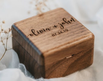 May include: A wooden ring box with a hinged lid. The top of the box is engraved with the text "Olivia + John" and the date "07.24.19".