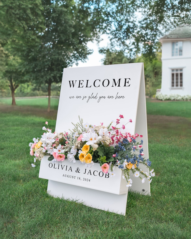 May include: White wooden welcome sign with a floral arrangement in a planter box. The sign reads "WELCOME we are so glad you are here OLIVIA & JACOB AUGUST 18, 2024".