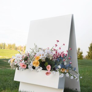 May include: A white A-frame sign with a blank white surface and a white planter box filled with a variety of colorful flowers. The sign is standing on a green grassy lawn.