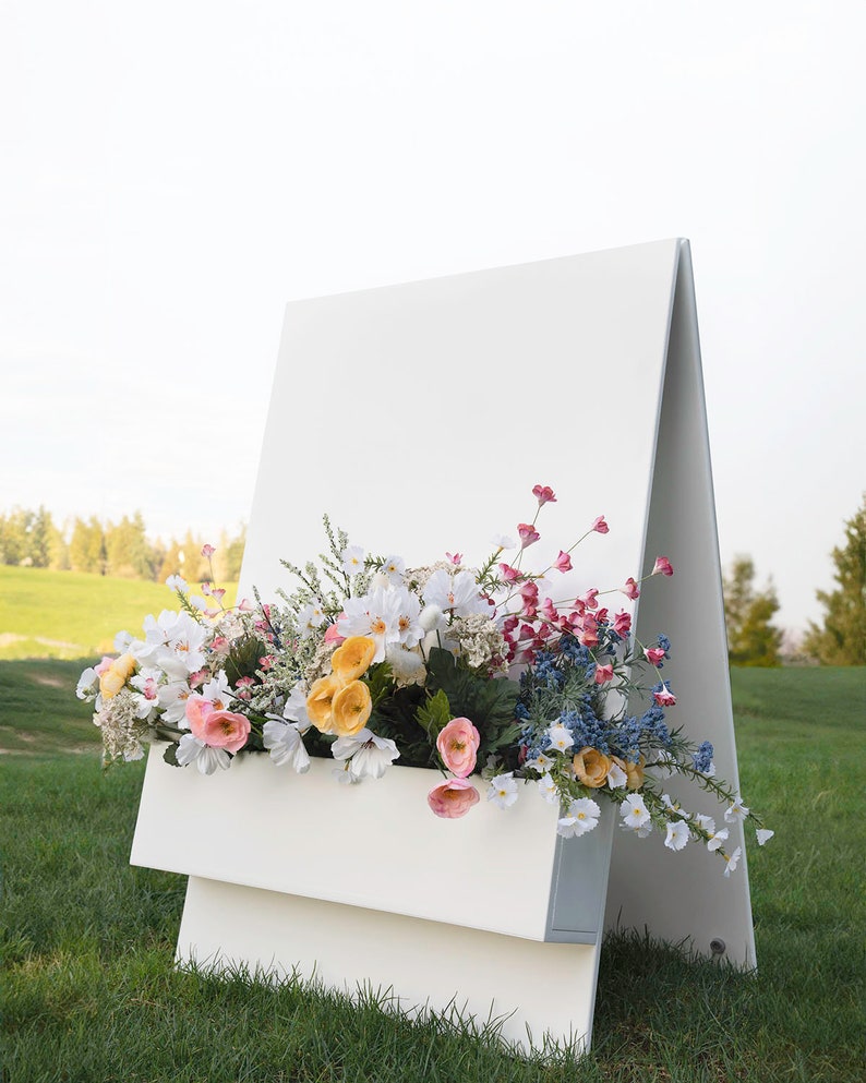 May include: A white A-frame sign with a blank white surface and a white planter box filled with a variety of colorful flowers. The sign is standing on a green grassy lawn.