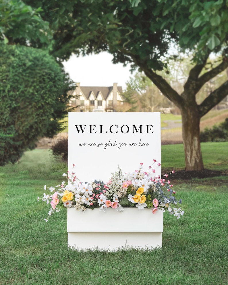May include: White welcome sign with the text "WELCOME we are so glad you are here" in black lettering. The sign is decorated with a planter box filled with pink, white, and yellow flowers.