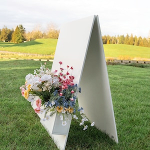 May include: A white A-frame sign with a built-in planter box filled with a colorful bouquet of flowers. The sign is standing on a grassy lawn.