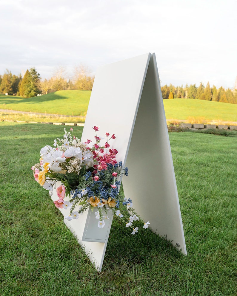 May include: A white A-frame sign with a built-in planter box filled with a colorful bouquet of flowers. The sign is standing on a grassy lawn.