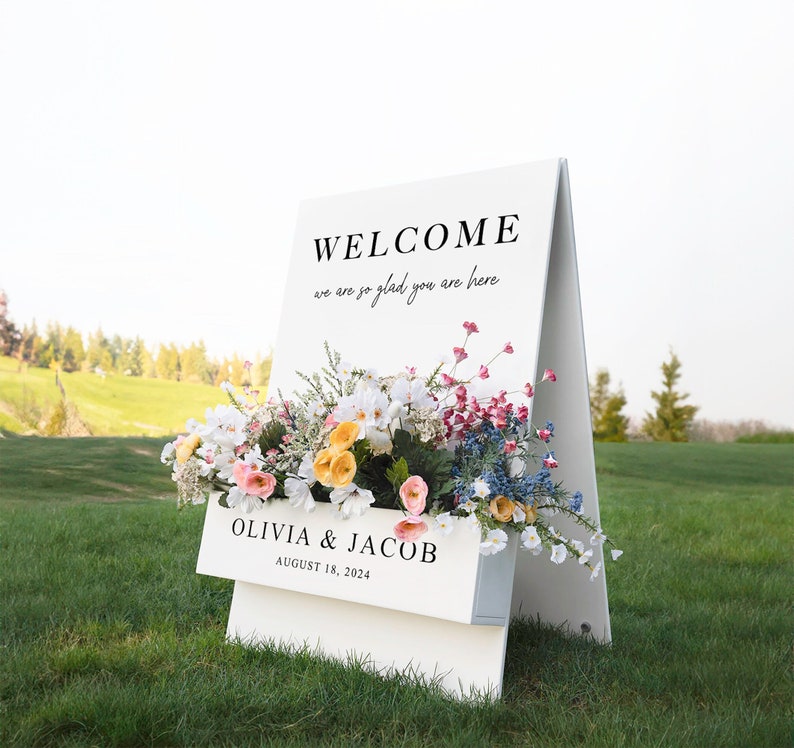 May include: White wooden A-frame sign with the text "WELCOME we are so glad you are here" and "OLIVIA & JACOB AUGUST 18, 2024". The sign is decorated with a floral arrangement of pink, yellow, white, and blue flowers.