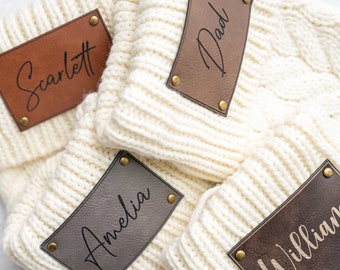 Puede incluir: Cuatro gorros de punto blanco con parches de cuero marrón. Los parches tienen los nombres "Dad", "Scarlett", "Amelia" y "William" escritos en cursiva.