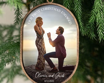 Op de afbeelding: Ovale houten kerstversiering met een foto van een huwelijksaanzoek. De tekst "OUR FIRST CHRISTMAS ENGAGED" staat bovenaan in een boog, met de namen "Olivia and Jacob" en de datum "8.12.2025" onderaan. Een wit lint is bevestigd.
