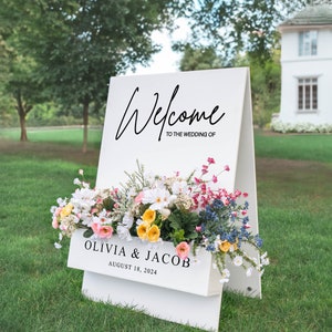 Puede incluir: Se&ntilde;al de bienvenida de boda blanca con un arreglo floral en una jardinera. El letrero dice "Bienvenido a la boda de Olivia & Jacob 18 de agosto de 2024".