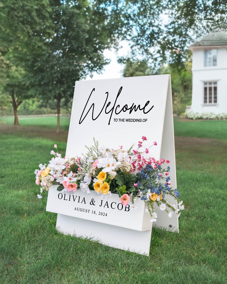 Puede incluir: Se&ntilde;al de bienvenida de boda blanca con un arreglo floral en una jardinera. El letrero dice "Bienvenido a la boda de Olivia & Jacob 18 de agosto de 2024".