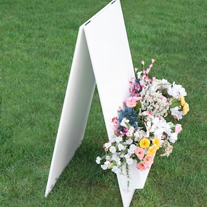 May include: A white A-frame sign with a blank white surface. The sign is decorated with a bouquet of pink, white, and yellow flowers.