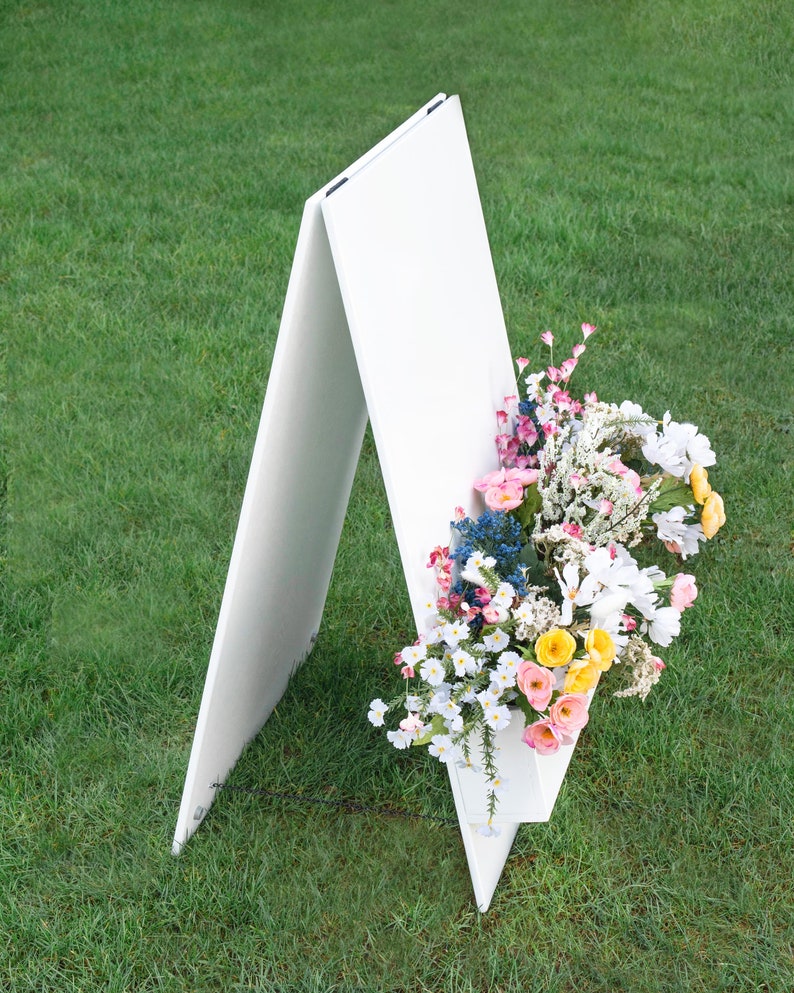 May include: A white A-frame sign with a blank white surface. The sign is decorated with a bouquet of pink, white, and yellow flowers.