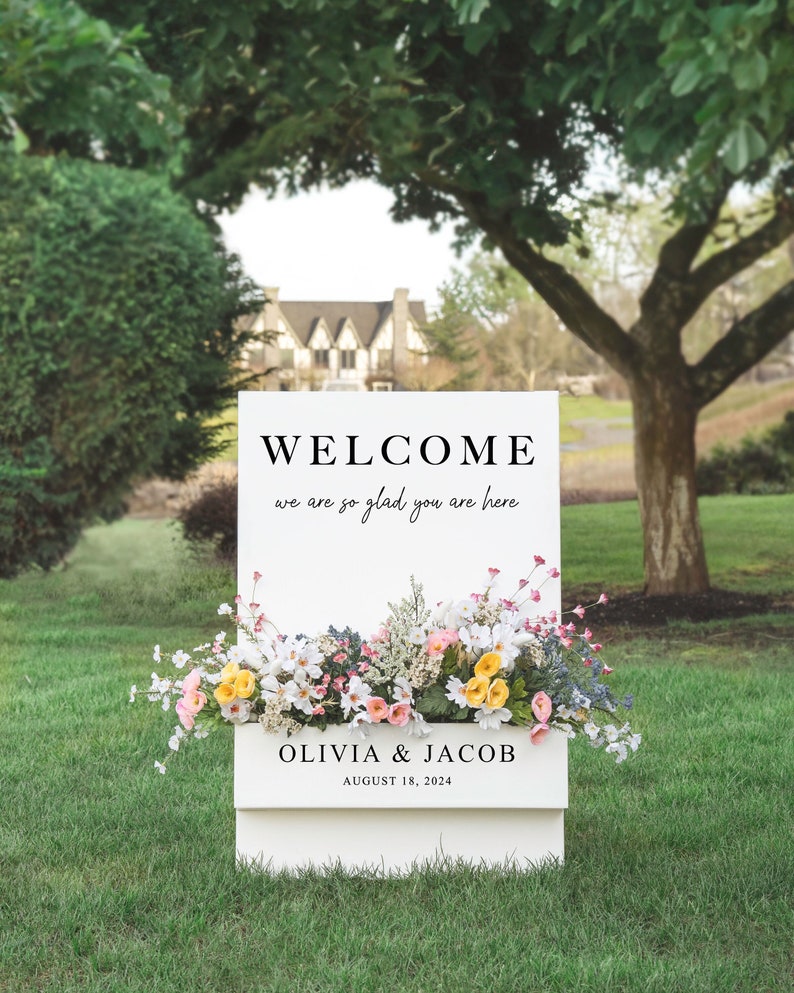 May include: White welcome sign with floral decorations, featuring the text "WELCOME we are so glad you are here" and "OLIVIA & JACOB AUGUST 18, 2024".