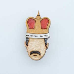 Könnte beinhalten: Holzbroschen-Anstecker mit einem stilisierten Porträt eines Mannes mit Schnurrbart und Krone. Die Krone ist goldfarben mit roten Akzenten. Die Brosche ist aus hellem Holz mit dunkelbraunen Details gefertigt.