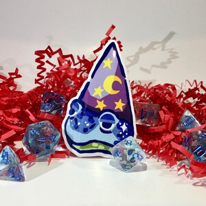 May include: A blue and purple wizard frog sticker with a crescent moon and stars on the hat. The sticker is surrounded by clear blue dice with white numbers.