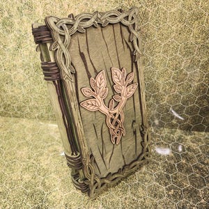 May include: A wooden journal with a leafy design carved into the cover. The cover is a light brown color with a green stain. The journal is closed and has a decorative metal clasp.