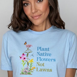May include: A light blue t-shirt featuring a graphic of a variety of flowers and the text "Plant Native Flowers Not Lawns".