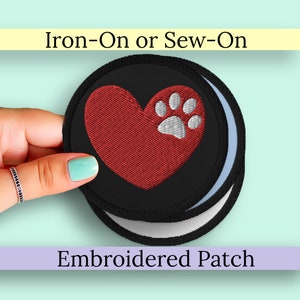 May include: Iron-on or sew-on embroidered patch with a red heart and a gray paw print on a black background. The patch is held by a hand.