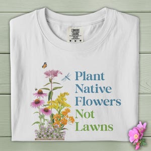May include: White t-shirt with the text "Plant Native Flowers Not Lawns" and a colorful floral illustration. The shirt features a Comfort Colors tag at the neckline. A butterfly and dragonfly are also depicted.