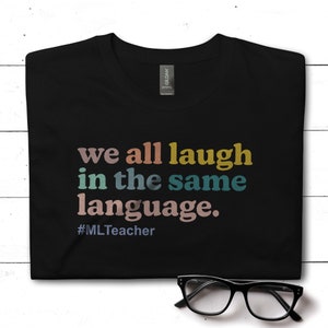 May include: Black t-shirt with the text "we all laugh in the same language." in rainbow colors and the hashtag "#MLTeacher" below.