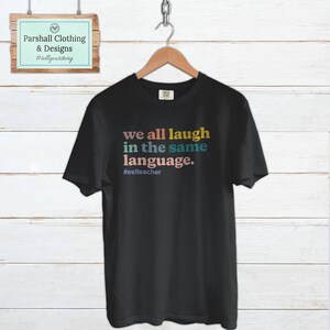 May include: Black t-shirt with the text "we all laugh in the same language." in rainbow colors. The text is followed by the hashtag "#eslteacher".