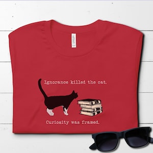 May include: Red t-shirt with a black cat and a stack of books. The text on the shirt reads "Ignorance killed the cat. Curiosity was framed."