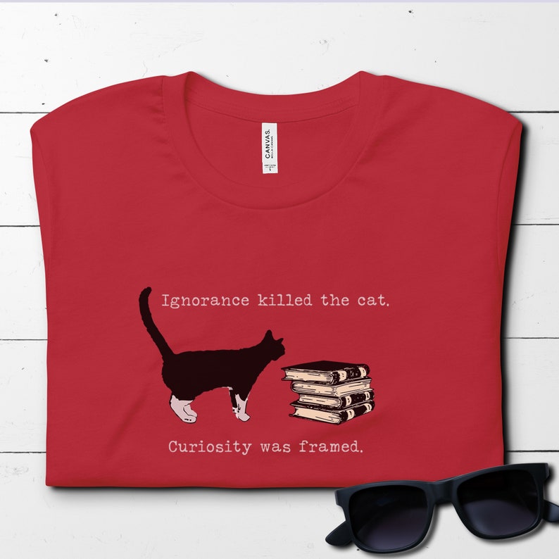 May include: Red t-shirt with a black cat and a stack of books. The text on the shirt reads "Ignorance killed the cat. Curiosity was framed."