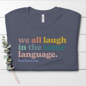 May include: A dark blue t-shirt with the text "we all laugh in the same language." in rainbow colors. The text is followed by the hashtag "#eslteacher".