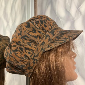 May include: A brown and black patterned hat with a short brim. The hat has a series of small, dark-colored studs along the top and a strap detail. The fabric has a mottled design.