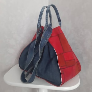 May include: A handmade tote bag with a patchwork design. The bag features a vibrant red body with denim accents, including the handles and interior lining. The red fabric is composed of various rectangular patches. The bag is sitting on a white surface.