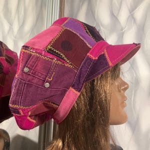Puede incluir: Una gorra de patchwork confeccionada con diversos paneles de tela en tonos rosa y morado. La prenda presenta costuras visibles en zigzag de color amarillo a lo largo de las uniones, remaches metálicos y una visera rígida y texturizada.
