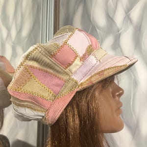 Puede incluir: Una gorra de estilo newsboy hecha de parches con diversas texturas y colores de tela, incluyendo paneles en rosa claro, beige y blanco. Las piezas están unidas con costuras en zigzag visibles y cuenta con una visera estructurada.