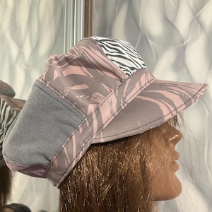Puede incluir: Un sombrero elegante con un diseño único. El sombrero presenta un ala gris y una corona con un estampado floral rosa y gris, acentuado por un panel con estampado de cebra. Un accesorio de moda.