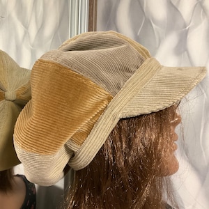 Puede incluir: Un sombrero de pana beige con un parche de pana marrón en el lateral. El sombrero tiene una ala ancha y una corona ligeramente curvada.