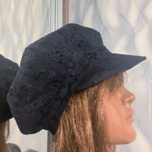 Peut inclure: Une casquette noire ornée d'un motif floral et cachemire ton sur ton sur tout le tissu. Ce couvre-chef présente une calotte structurée et une visière rigide horizontale s'étendant vers l'avant, affichant un design sombre et monochrome.