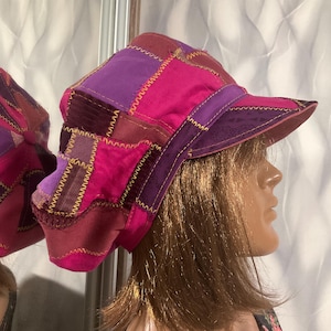 May include: A patchwork newsboy cap featuring various shades of pink and purple fabric panels. The hat is constructed with visible yellow zigzag stitching connecting the different textile pieces and includes a stiffened brim in a dark purple tone.