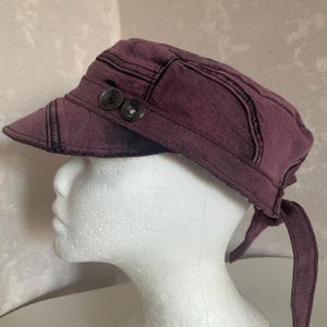 May include: A dark purple cadet cap with a distressed, washed-out finish. The hat features a short, stiff brim and two dark circular buttons attached to the side. It includes fabric ties at the back for adjustment, providing a secure fit.