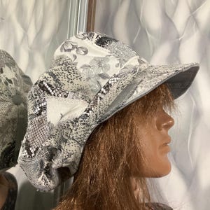 May include: A newsboy cap featuring a patchwork design of various fabrics in shades of gray, white, and black. The hat displays a mix of floral patterns, snake skin textures, and solid panels with visible stitching along the seams and a stiff brim.