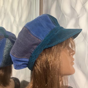 Puede incluir: Una gorra de estilo newsboy azul patchwork con un borde de pana turquesa. La gorra presenta paneles de tela vaquera y de imitación de ante, cosidos con hilo en contraste. El borde es de color turquesa liso. La gorra se muestra en un maniquí.