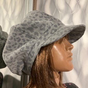 May include: A gray newsboy cap with a leopard print pattern. The hat has a rounded crown and a short, stiff brim. The fabric appears to be a soft, woven material. The cap is displayed on a mannequin head.