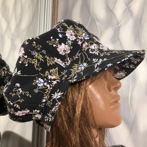 May include: A black hat with a floral pattern featuring pink, blue, and green flowers. The hat has a wide brim and a structured design, suitable for sun protection. The floral print adds a touch of elegance to the hat.