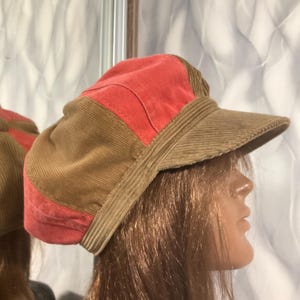 May include: A corduroy newsboy cap in shades of rust and brown. The cap features a structured design with a short brim and a panel of rust-colored fabric on top. The hat is made of a soft, textured corduroy material.