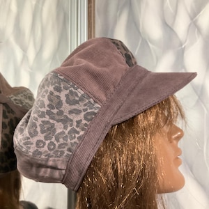 Puede incluir: Un sombrero de pana malva con un estampado de leopardo. El sombrero presenta una visera estructurada y un panel de tela con estampado de leopardo en el lateral. El sombrero está diseñado con un estilo clásico.