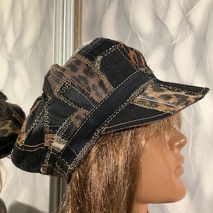 Puede incluir: Una gorra de estilo newsboy de patchwork que combina paneles de tela negra y secciones con estampado de leopardo marrón. La gorra presenta costuras en zigzag visibles con hilo dorado a lo largo de las uniones y una visera rígida.