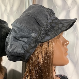 May include: A dark gray newsboy cap with a patterned brim and a camouflage design on the crown. The cap has a short, stiff brim and a rounded top. The fabric appears to be a soft, durable material.