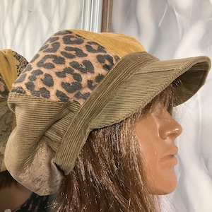 Puede incluir: Una gorra de newsboy de pana marrón con un panel de estampado de leopardo. La gorra tiene una visera ancha y una sensación suave y cómoda.