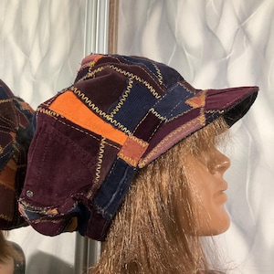 Puede incluir: Una gorra de estilo newsboy hecha de parches con diversas texturas de tela, incluyendo pana y terciopelo en tonos morado oscuro, azul marino y naranja brillante, con costuras en zigzag amarillas a lo largo de las uniones y una visera rígida.