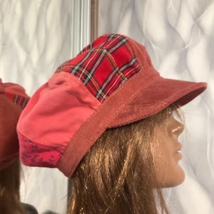 Puede incluir: Una gorra estilo newsboy de múltiples texturas con un panel de tela de cuadros rojos y negros, una sección de algodón rosa coral sólido y una banda inferior con estampado rosa. La gorra incluye una visera estructurada de pana coral.