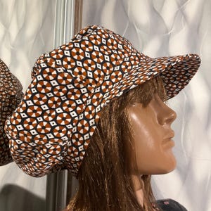 May include: A patterned hat with a brim, featuring a repeating geometric design in brown, black, and white. The hat is worn by a mannequin. The hat's design includes diamond and circular shapes.
