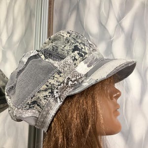 May include: A gray and white patchwork cap featuring a mix of denim and snakeskin-patterned fabric panels. The hat is constructed with visible white decorative stitching along the seams and includes a structured brim and a rounded crown design.