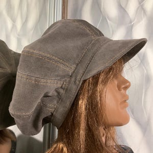 Puede incluir: Una gorra de estilo newsboy en pana gris con una construcción de múltiples paneles y costuras visibles en color tostado a lo largo de las uniones. El gorro presenta una visera estructurada y una superficie de tela texturizada con silueta redondeada.